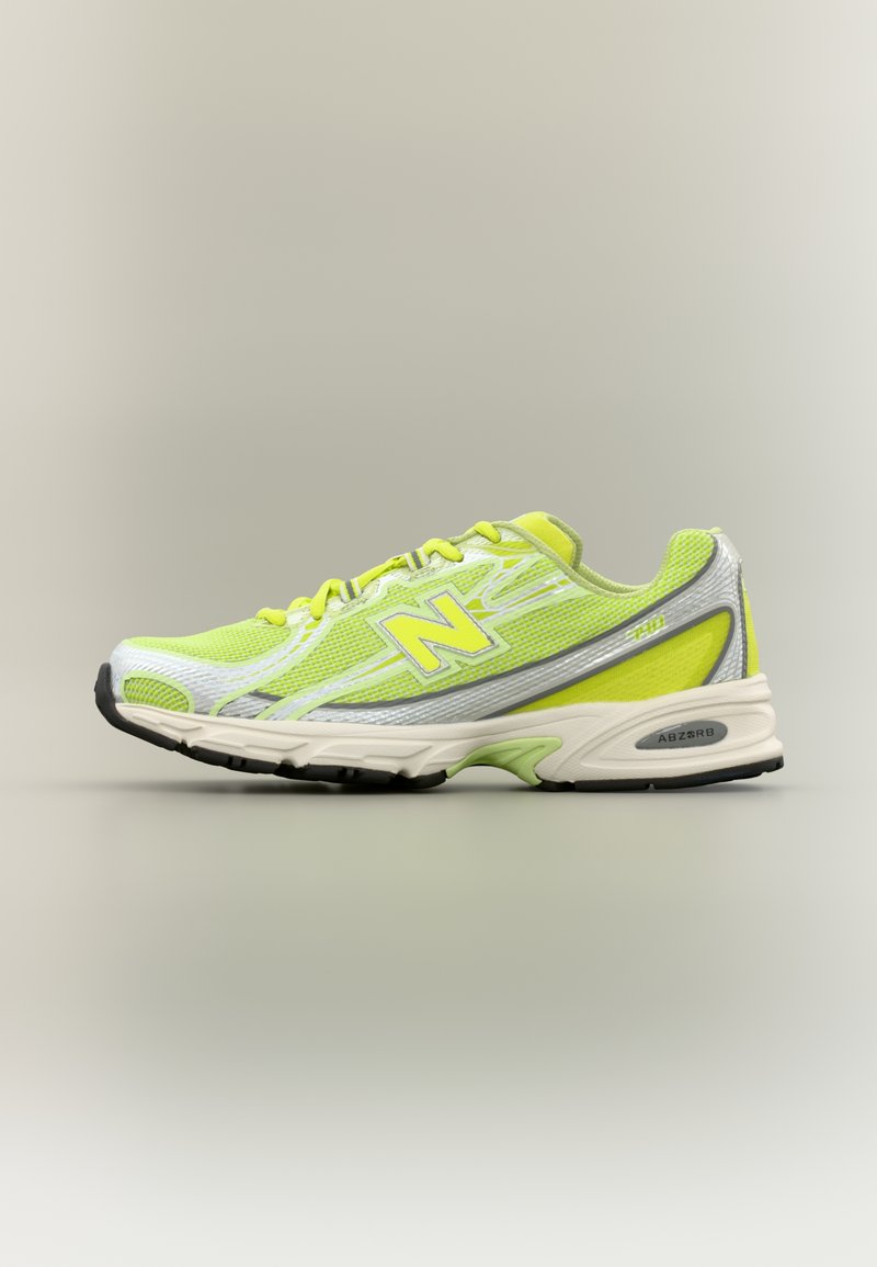 Αθλητικό παπούτσι New Balance σε ανοιχτό πράσινο και ασημί με κίτρινες λεπτομέρειες, λευκή σόλα και μαύρο πέλμα, σε πλάγια προβολή σε ουδέτερο φόντο.