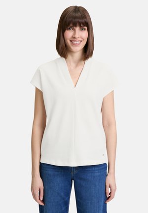 Top bianco a maniche corte con scollo a V. Il design presenta una vestibilità comoda e un piccolo logo a rilievo sull'orlo. Indossato con jeans blu.