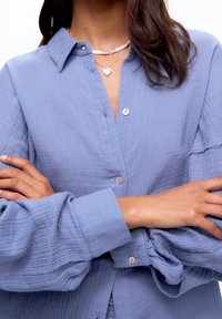 Ljusblå, texturerad blus med knäppning framtill, långa voluminösa ärmar och krage. Har en subtil rynkad design.