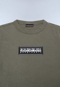 Olivenfarbene Baumwoll-T-Shirt mit Rundhalsausschnitt und einem rechteckigen schwarzen Grafiklabel, das "NAPAPIJRI geographic" in weißem Text zeigt.