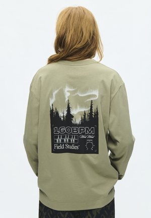 Persona con i capelli lunghi indossa una maglietta verde a maniche lunghe con una grafica nera di alberi, fumo bianco e testo con scritto "160 BPM Field Studies."