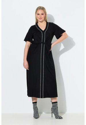 Kvinde iført en sort midikjole med hvide lodrette striber og knudedetalje, kombineret med sorte og hvide mønstrede ankelstøvler med høje hæle.