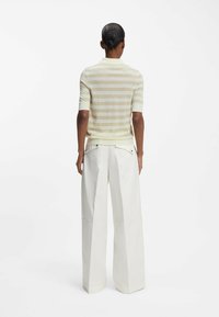 Persona vista da dietro che indossa una maglietta a maniche corte beige a righe e pantaloni bianchi a vita alta e larghi, su uno sfondo bianco.