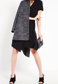 Musta mekko, jossa on epäsymmetrinen helma, kerroksittain harmaalla tekstuuritakilla. Mallella on pieni musta laukku ja mustat avokkaat.