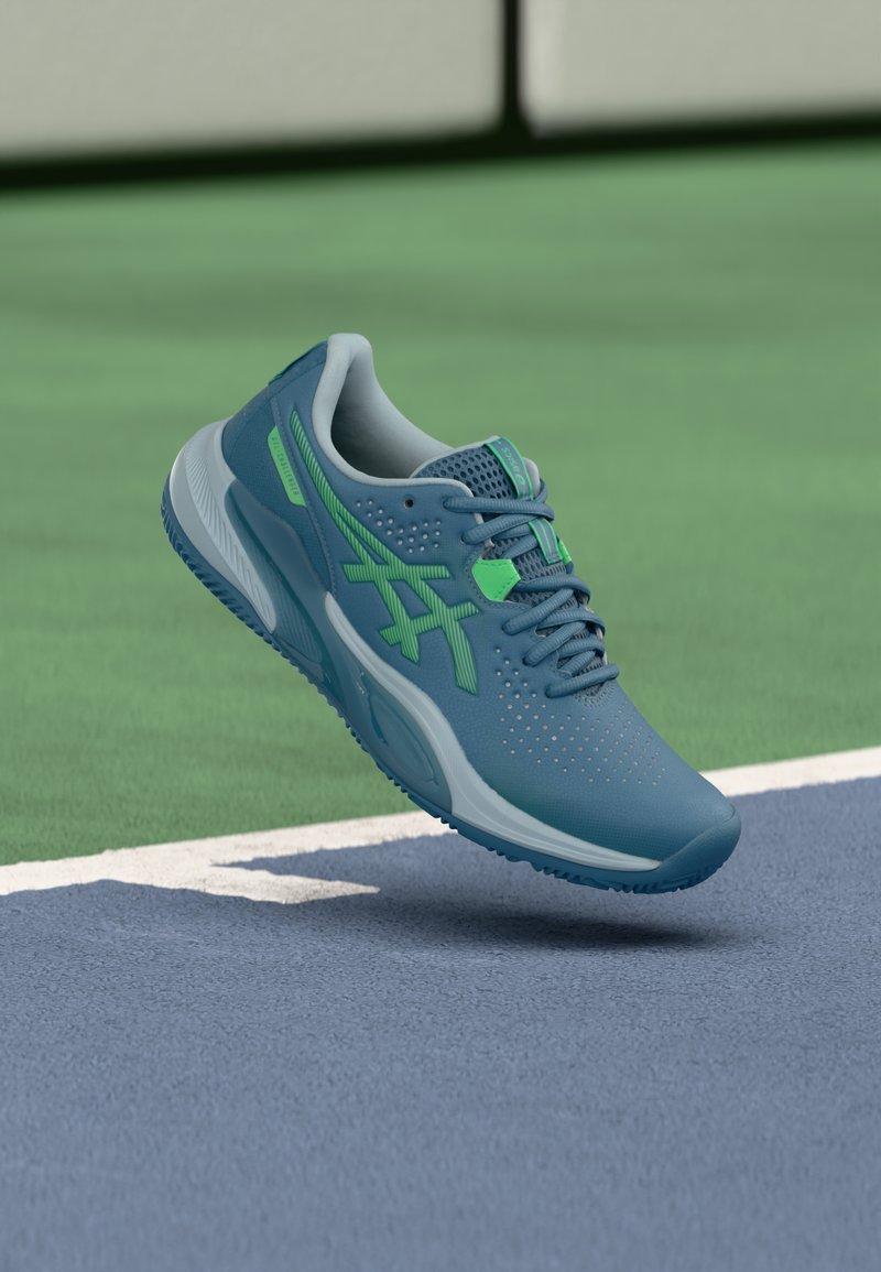 Scarpa da ginnastica blu con dettagli verdi, tomaia in mesh, suola testurizzata, fori per la traspirazione e design leggero.