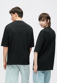 Μαύρο υπερμεγέθες μπλουζάκι με κοντά μανίκια, λαιμόκοψη τύπου crew neck, κατασκευασμένο από μαλακό βαμβάκι, σε συνδυασμό με φαρδιά τζιν σε ανοιχτό μπλε χρώμα, εμφανιζόμενα από πίσω.