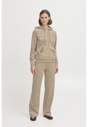 Vrouw staand, gekleed in een beige capuchonvest met ritssluiting en bijpassende broek, met zwarte balletschoenen, tegen een effen lichte achtergrond.