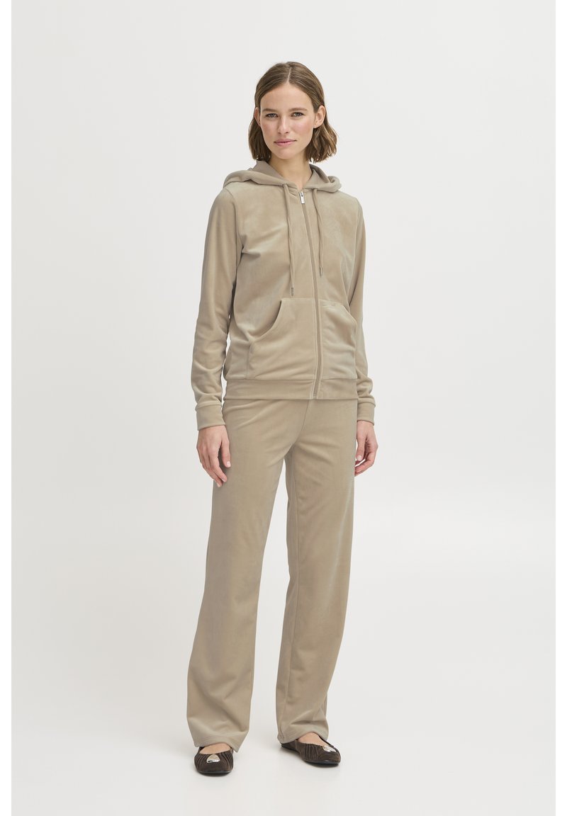 Naine seisab seljas beež värvi lukuga kapuutsiga jakk ja sobivad püksid ning mustad baleriinikingad, taust on hele ja ühtlane.