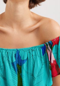 Žena v barevném topu s odhalenými rameny, na kterém jsou vyobrazeny modré, zelené, červené a růžové květinové vzory, který odhaluje výstřih a klíční kosti.