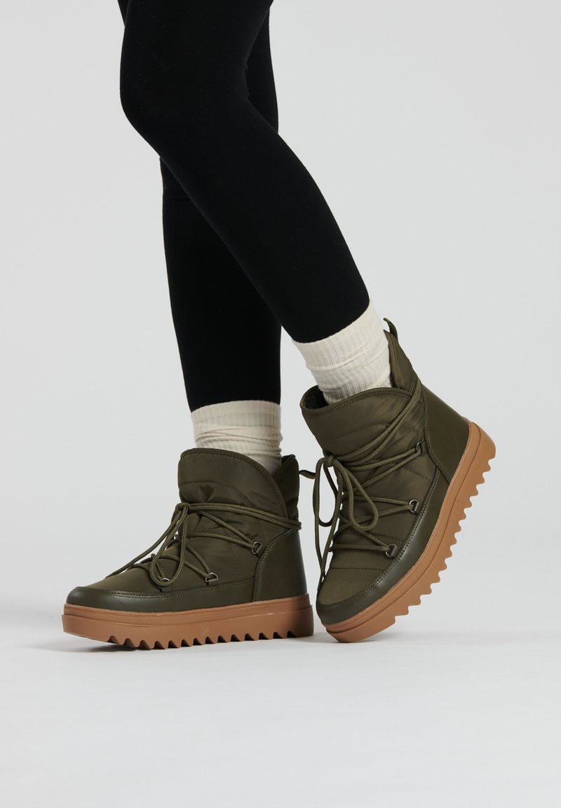 Bottes de cheville vertes imperméables avec un col rembourré, un laçage à l'avant et une épaisse semelle brune texturée, associées à des chaussettes blanches côtelées et des leggings noirs.