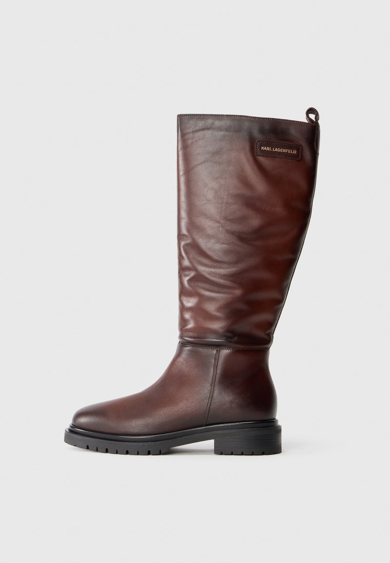 Barna bőr, térd fölé érő csizmák, enyhén megereszkedett dizájnnal, kerek orral és vastag, texturált fekete gumitalppal. Logópatent található rajtuk.