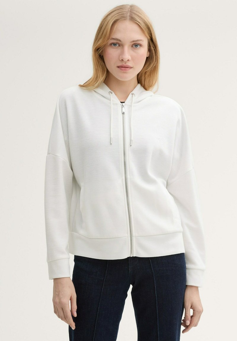 Sweat à capuche zippé blanc avec capuche, poignets et ourlet côtelés. Texture lisse avec cordons de serrage et poches latérales cachées. Design ajusté.