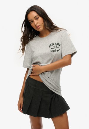 T-shirt en coton gris avec le logo "Superdry" en noir, associé à une jupe plissée à carreaux noirs, mettant en valeur une coupe décontractée.
