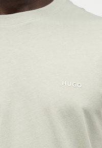 Κοντινό πλάνο ενός ανοιχτόμπεζ μπλουζάκι με μικρό λευκό λογότυπο «HUGO» στο στήθος, που φοριέται από άτομο με σκούρο δέρμα.