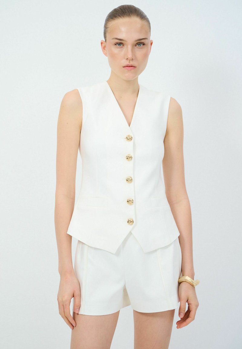 Chaleco blanco sin mangas con escote en V, adornado con seis botones dorados y bolsillos laterales, combinado con pantalones cortos blancos a medida. Textura suave.