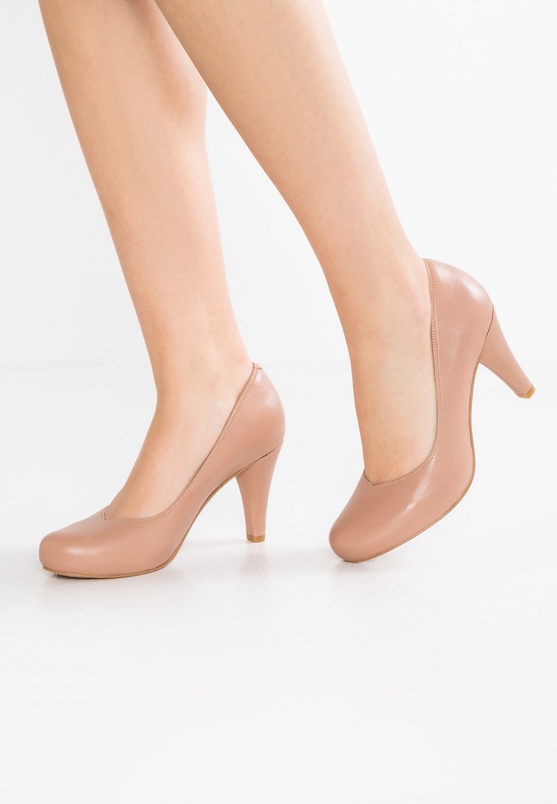 Femme portant des escarpins en cuir beige à talons moyens, debout sur une surface blanche avec un pied légèrement levé.