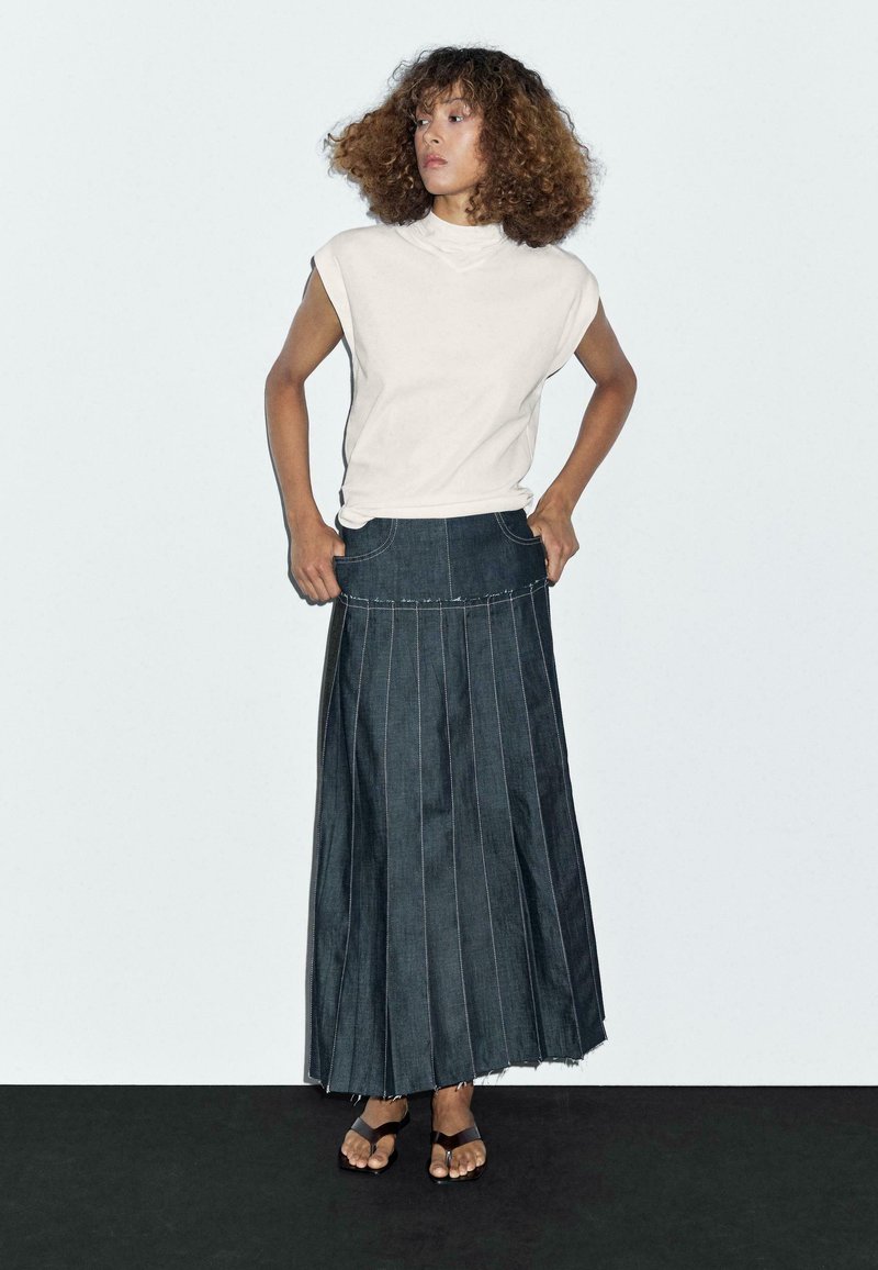 Witte mock neck mouwloze top gecombineerd met een donkerdenim maxi rok met verticale stiksels en een rafelige zoom. Bruine sandalen.