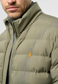 Muškarac nosi maslinasto zeleni kaput s prešivom s malim vezom narančaste figure na prsima, djelomično zatvoren preko sive turtleneck dukserice.