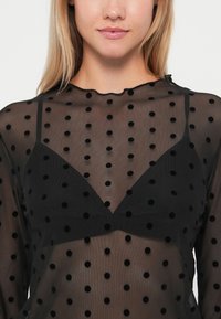 Zwarte doorzichtige top met ronde fluwelen stippen, met een scalloped halslijn en een zwarte bralette eronder.