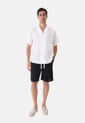 Uomo con camicia bianca a maniche corte abbottonata, pantaloncini neri con coulisse e scarpe beige senza lacci, in piedi con una mano in tasca.
