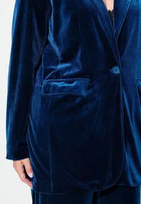 Sininen samettitakki, jossa on pehmeä rakenne, kahdella etutaskulla, yhdellä napilla kiinnitys ja istuva malli.