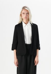 Svart blazer med skreddersydd passform, litt forkortede ermer og en jevn tekstur. Kombinert med en hvit skjorte med knapp.