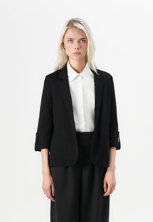 Black blazer with a tailored fit, slightly cropped sleeves, and a smooth texture. Paired with a white, button-up shirt.