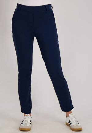 Marineblauwe slim-fit broek gecombineerd met witte sneakers met zwarte strepen en beige neuzen, gedragen door een staande persoon tegen een effen achtergrond.