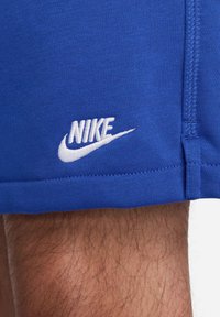 Blå Nike-shorts av stretchigt tyg; har en vit broderad logotyp nära fållen; slät textur och rak siluett.