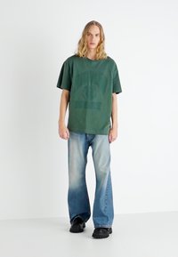 Camiseta verde oversized con un logo impreso, combinada con jeans acampanados de color azul claro y zapatos gruesos negros, de pie frente a un fondo blanco.
