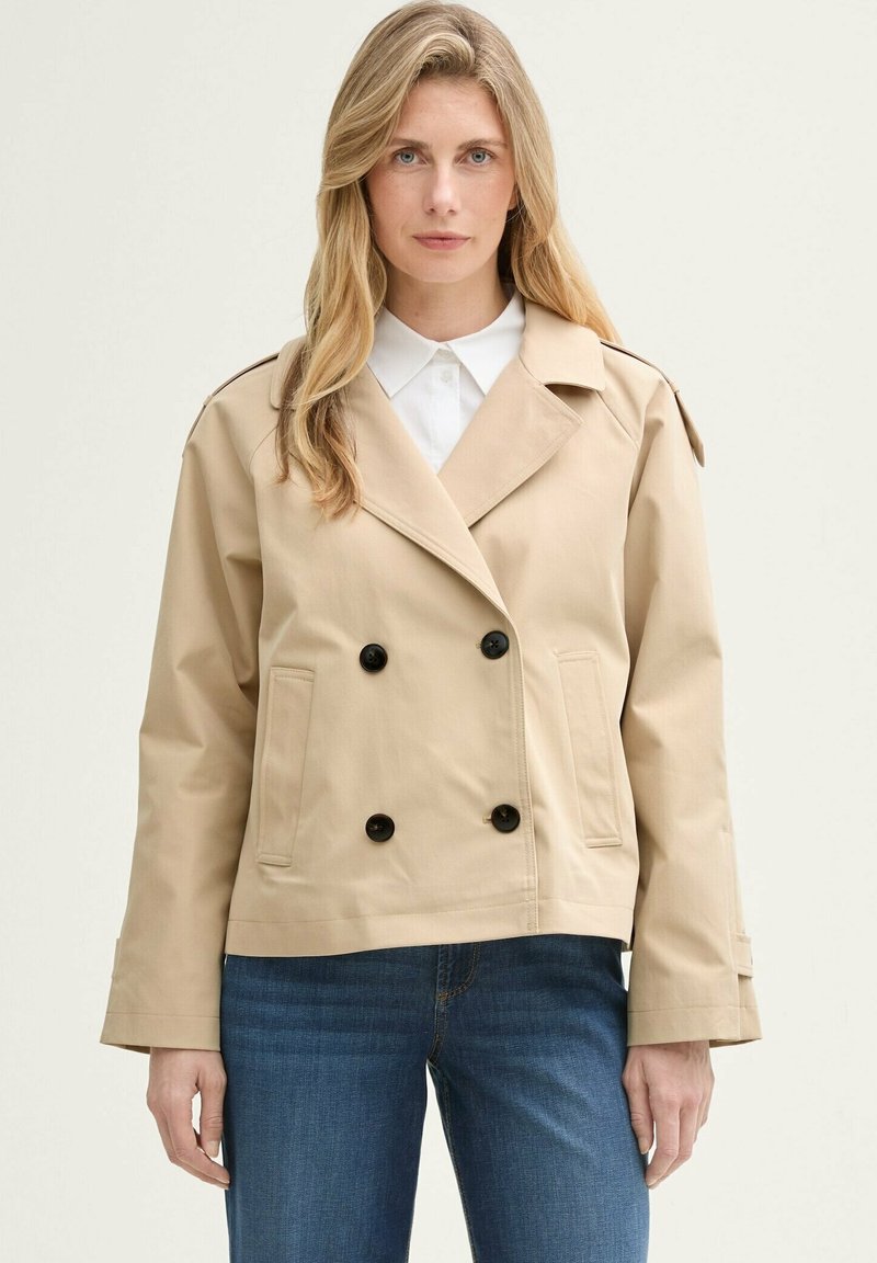 Beige Doppelreiher-Jacke aus glattem Stoff, mit schwarzen Knöpfen, einem aufgeschlagenen Kragen und Seitentaschen, getragen über blauen Jeans.