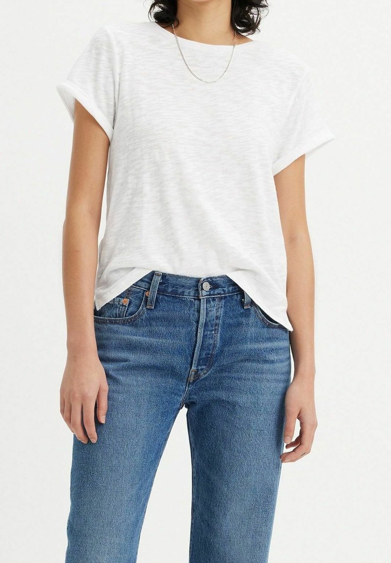 Femme portant un t-shirt blanc uni à manches courtes, légèrement rentré dans un jean bleu taille haute avec un collier chaîne argenté.