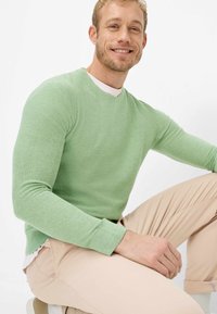 Lichtgroene gebreide trui met een ronde hals en lange mouwen, gedragen met licht beige broek. De stof heeft een gestructureerde uitstraling.