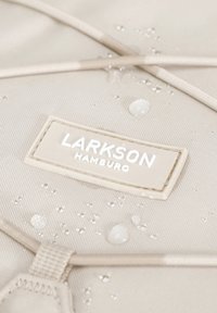 Beige vízhatlan szövet, texturált felülettel, Larkson Hamburg címkével és apró vízcseppekkel. Rugalmas pántokkal kiegészítve.