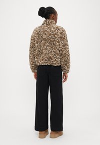 Vachtjas met een fuzzy bruin en crème patroon en een hoge kraag, gedragen over zwarte broek met wijde pijpen en tan kleurige schoenen, gezien van achteren.