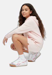 Abitino in felpa rosa chiaro con fessure laterali, abbinato a sneaker bianche con dettagli rosa e suola in gomma. La modella si accovaccia contro uno sfondo chiaro.