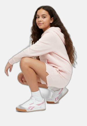 Vestido sudadera de color rosa claro con aberturas laterales, combinado con zapatillas blancas con acentos en rosa y suela de goma. La modelo está agachada contra un fondo claro.