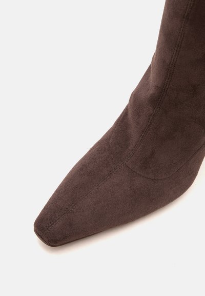 Gros plan d'une botte en daim marron à bout pointu avec des coutures visibles sur fond blanc.