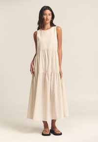 Beige ärmlös maxikjol med ett lagerdesign, rund halsringning och mjuk textur. Bärs med svarta plana flip-flops.