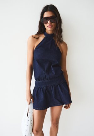 Ženska, oblečena v temno modro obleko s halterjem in velikimi sončnimi očali, drži belo torbico z netki na enobarvni bel ozadju.