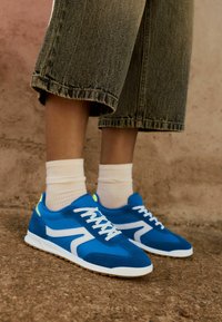 Baskets de sport bleues en suède et matériaux synthétiques, avec des accents blancs, une semelle en caoutchouc texturé, et portées avec des chaussettes beiges côtelées.