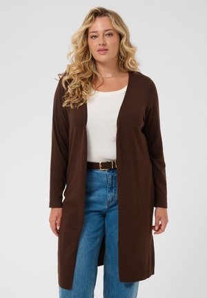 Femme blonde aux cheveux bouclés portant un long cardigan marron, un haut blanc, un jean bleu et une ceinture noire avec une boucle dorée.