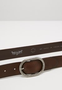 Ceinture en cuir marron avec une boucle ovale en argent, dotée de perforations, d'un logo en relief et de détails textuels sur la sangle intérieure et le verso.