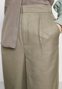 Béžové plisované kalhoty vyrobené z texturované tkaniny, s bočními kapsami a vysokým pasem, doplněné neutrálním topem.