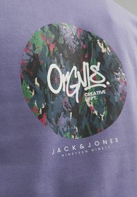 Camiseta de algodón morada con un gráfico circular que presenta un diseño abstracto colorido y texto blanco que dice "ORGNLS. CREATIVE DEPT." y el nombre de la marca.
