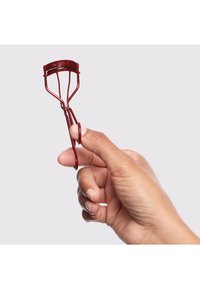 Piegaciglia in metallo rosso tenuto tra le dita, con cuscinetto in gomma curvo e impugnature arrotondate. Finitura liscia con un design minimalista.