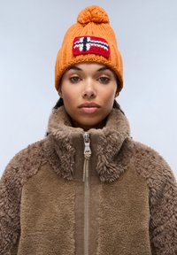 Pletená oranžová čepice s pomponem a pruhovanou nášivkou; hnědá fleecová bunda se zipem vpředu a texturovanými detaily na ramenech a límci.