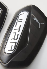 Zwarte voetbalkniebeschermers met een glanzend zilveren "ULTRA"-logo en Puma-logo, met een gladde oppervlakte en een gestroomlijnd, ergonomisch ontwerp.