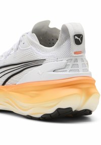 Zapatilla deportiva blanca con parte superior de malla, rayas negras, acento naranja y suela gruesa multicapa con diseño de olas.