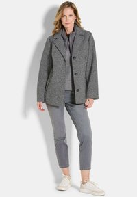 Frau, die einen grauen Fischgrätenmantel über einer gesteppte Jacke und grauen cropped Hosen trägt, mit weißen Sneakers vor einem weißen Hintergrund.